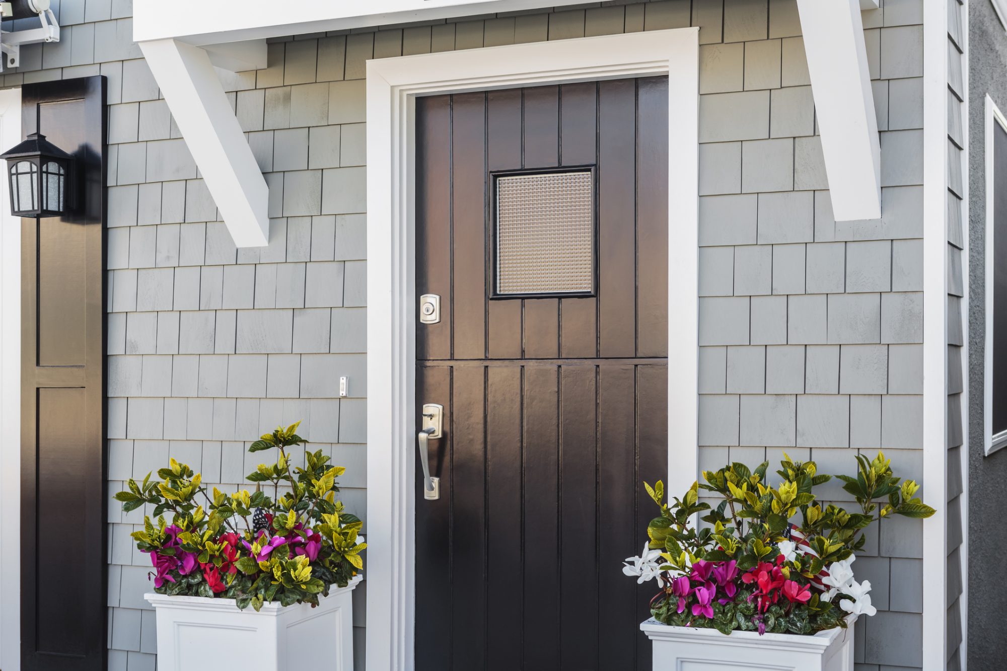 Angled view of glossy black front door to a family home; The door is made of vertical wood boards, with a window. It is framed by two flower planters, gray shingles of the house, and a door mat.